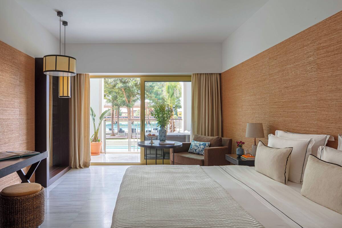Habitación de hotel con una cama grande, un escritorio, una silla, una mesita con un jarrón y vistas al exterior.