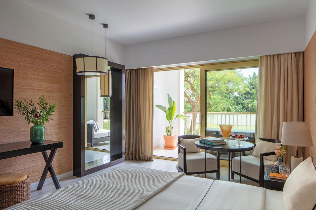 Habitación de hotel con cama, escritorio, silla, mesa y balcón con vistas a zonas verdes.