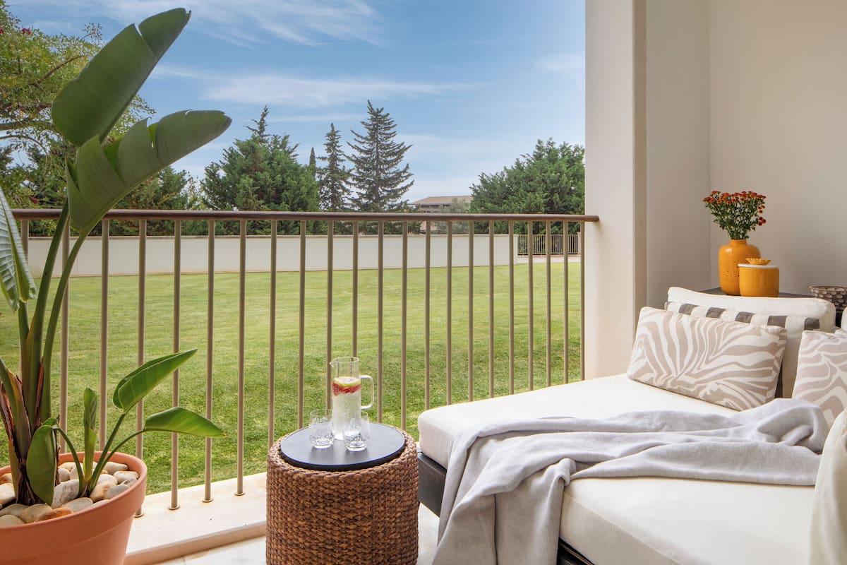 Un balcón con vistas a un área verde, una planta en maceta, una pequeña mesa redonda con una jarrón y vasos, y una cama con almohadas y una manta.