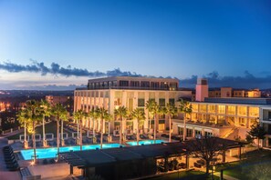 A hotel with a pool, palm trees, and a view of the city at dusk. - Victoria Golf Resort and Spa Managed by Accor (Loule)