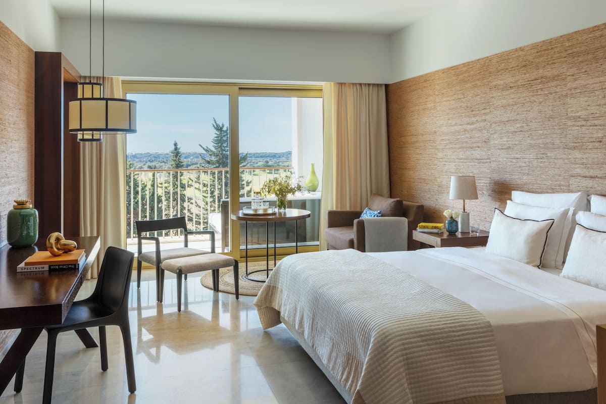 Habitación de hotel con una cama grande, un escritorio con silla, una mesita de noche con lámpara y vistas al exterior.