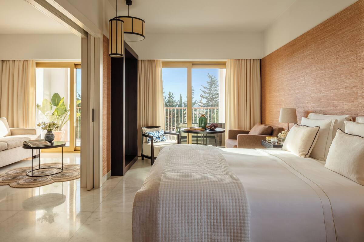 Una habitación de hotel moderna con una cama grande, un balcón con vistas, una zona de estar con una mesa y un ventanal amplio.