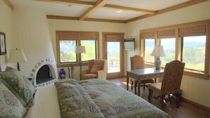 A room with a fireplace, a bed with patterned bedding, a table with a lamp, and large windows offering a view of the outdoors.