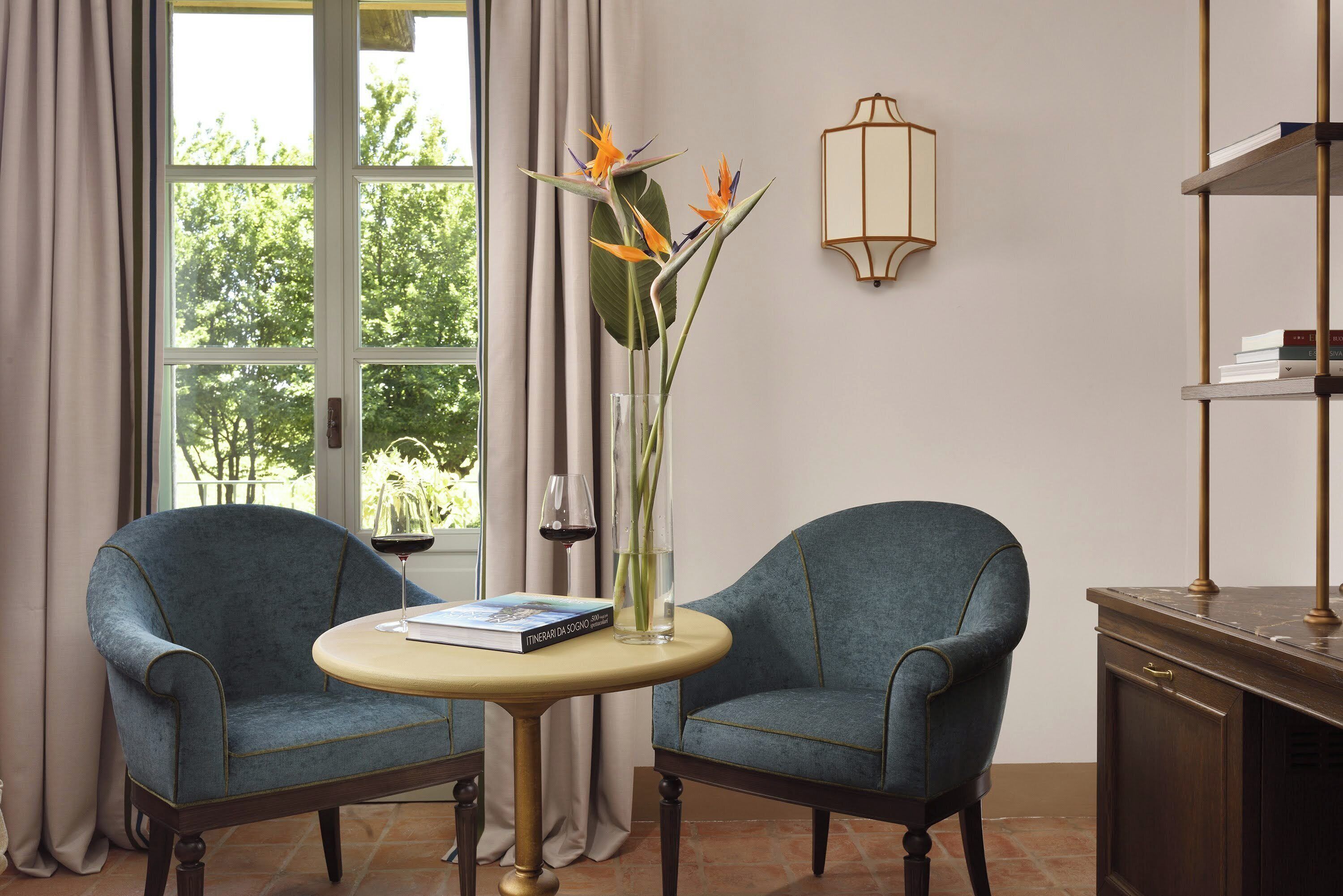 Una habitación con dos sillones, una mesita redonda, un jarrón con flores, una copa de vino y una estantería.