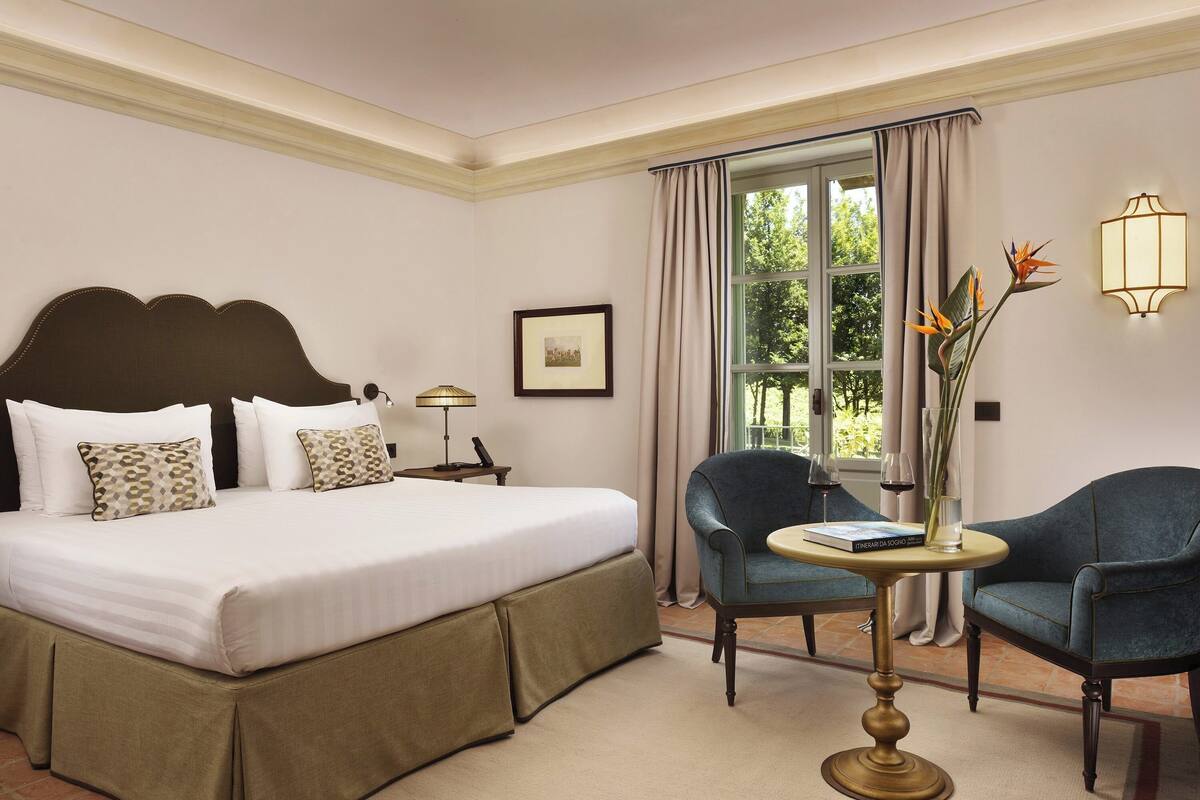 Une chambre d’hôtel avec un grand lit, deux fauteuils, une petite table, une lampe, un tableau encadré et une fenêtre avec des rideaux.