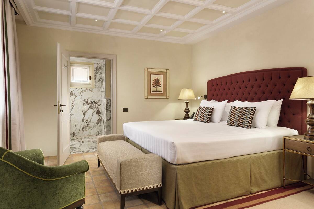 Une chambre avec un grand lit, un banc, une table de chevet, une lampe, un tableau encadré au mur et une porte avec une fenêtre.