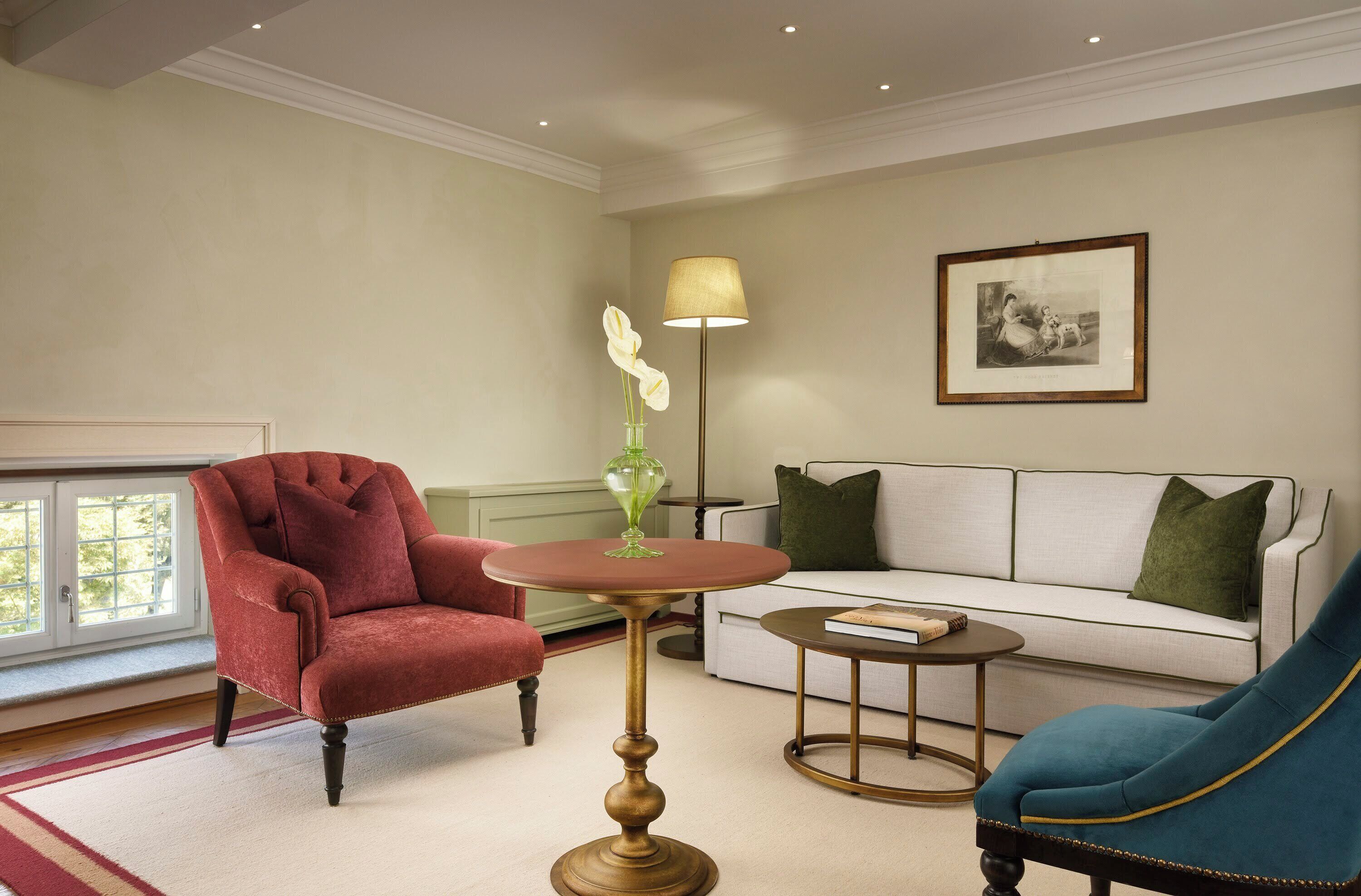 Ein Wohnzimmer mit einem roten Sessel, einem blauen Sessel, einem weißen Sofa mit grünen Kissen, einer Stehlampe und einem gerahmten Bild an der Wand.