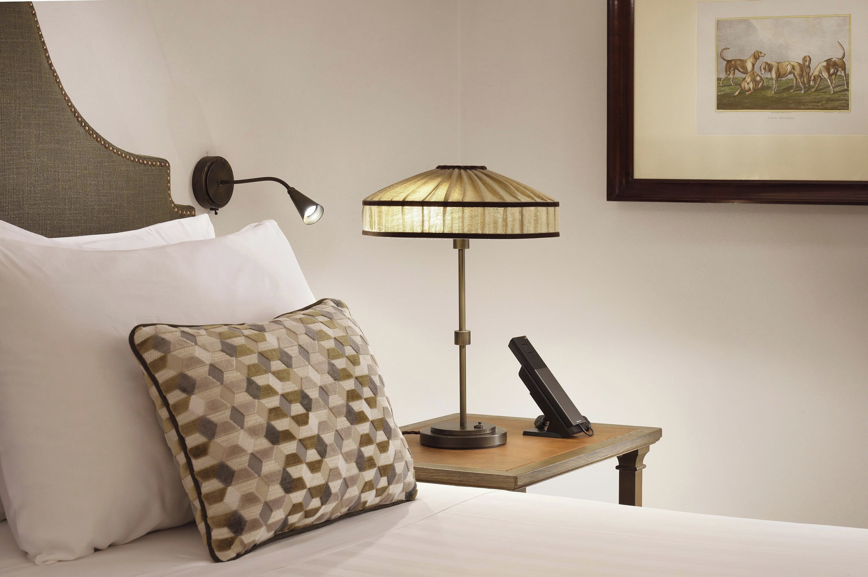 Ein Hotelzimmer mit einem Bett, einem gemusterten Kopfkissen, einer Wandlampe, einem kleinen Tisch mit Telefon und einem gerahmten Bild an der Wand.