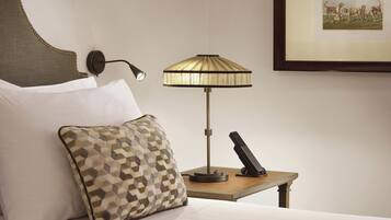 Una habitación de hotel con una cama, un cojín estampado, una lámpara de pared, una mesita con un teléfono y un cuadro enmarcado.