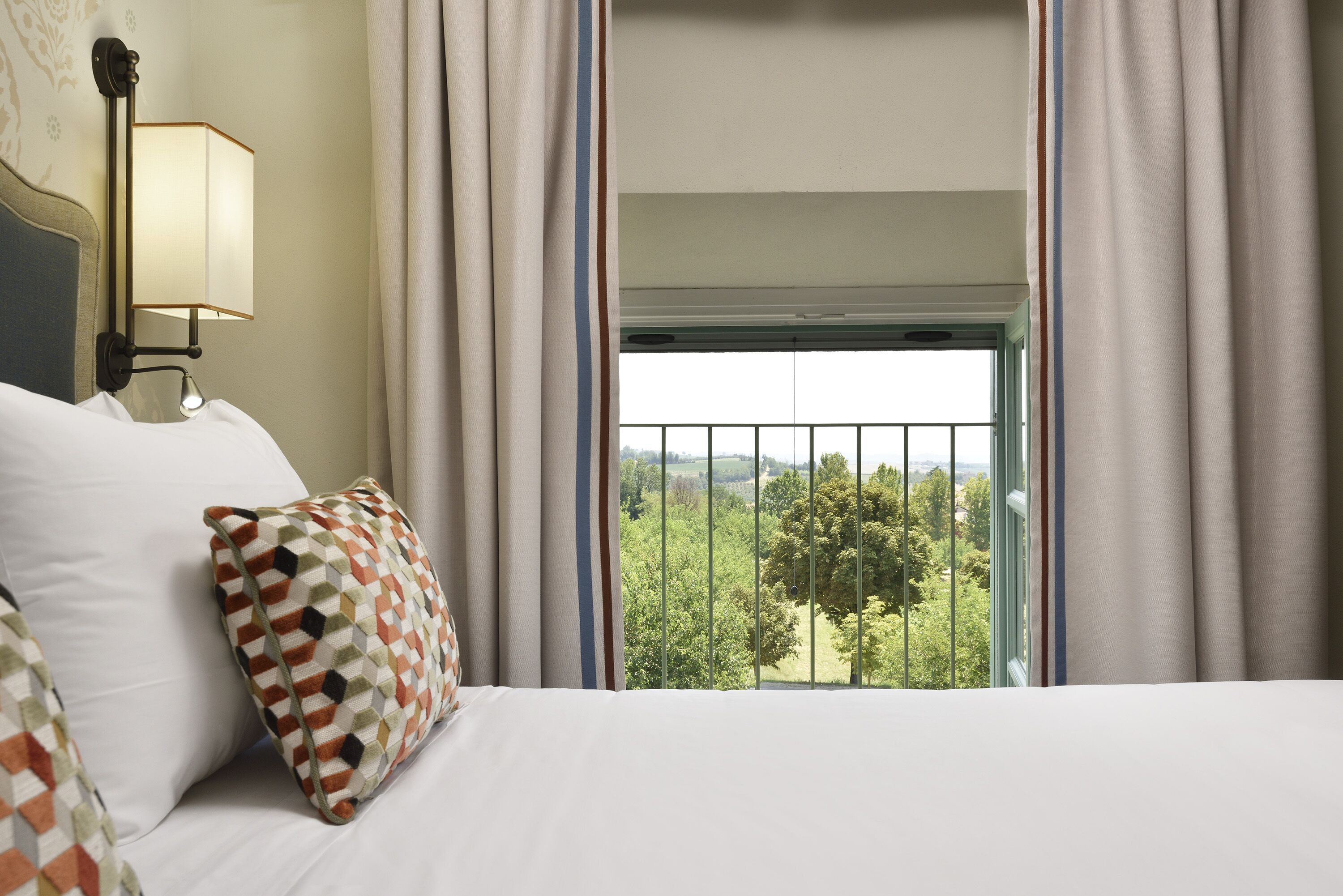 Une chambre d’hôtel avec un lit, un coussin à motifs, une table de chevet avec une lampe et une vue sur la verdure par une fenêtre avec des rideaux.