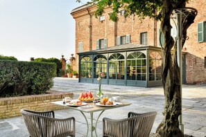 Un edificio de ladrillo con un invernadero de vidrio, asientos exteriores con sillas de mimbre y una mesa dispuesta con comida y bebidas.