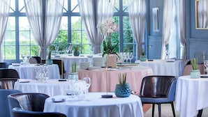 A dining area with tables set for service, large windows with curtains, and potted plants.