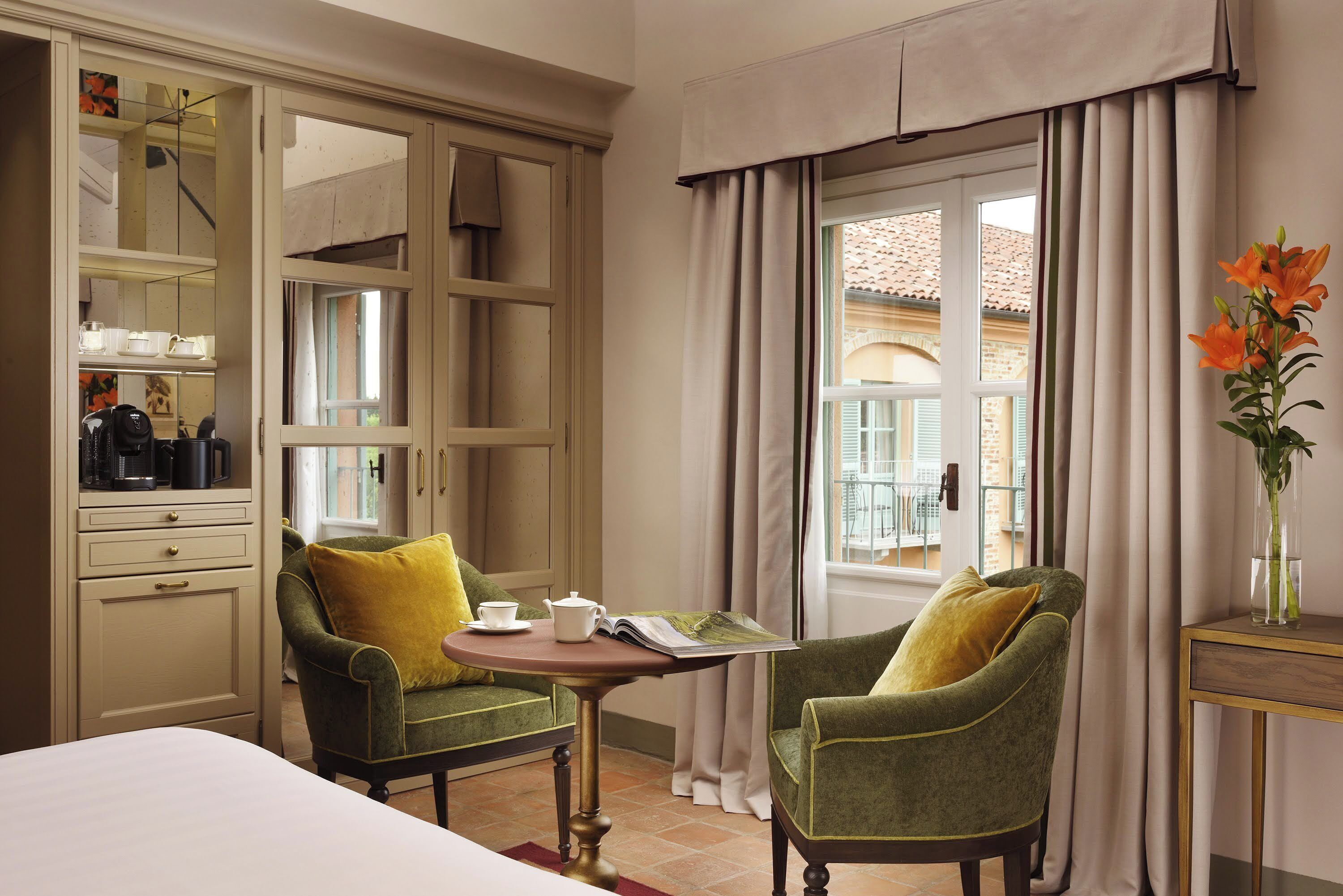 Ein Hotelzimmer mit einem Bett, zwei grünen Sesseln, einem kleinen Tisch mit einem Buch und einer Teekanne, einem Spiegel und einem Fenster mit Vorhängen.