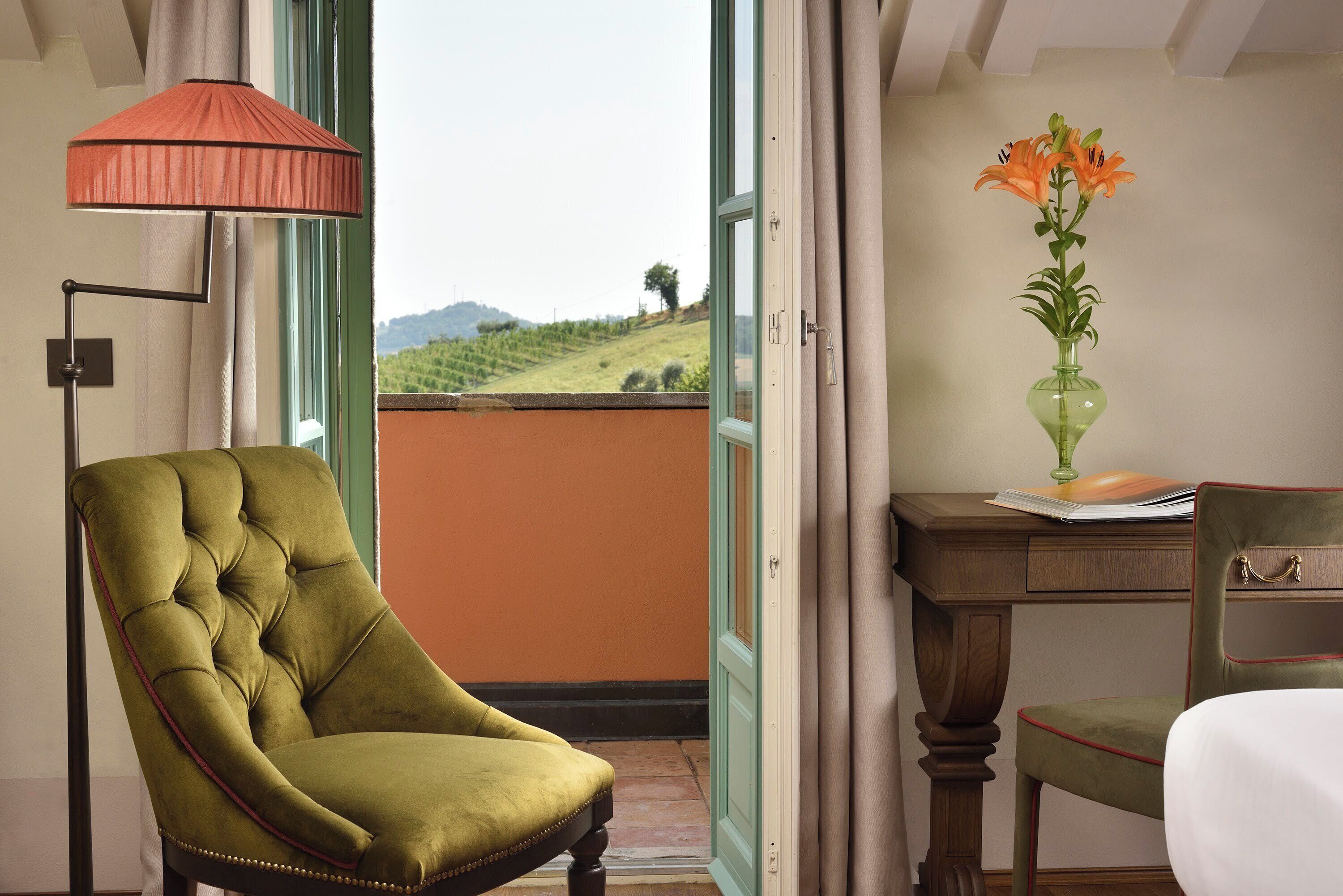 A room with a green armchair, a desk with a vase of flowers, and a view of a green landscape through a window.