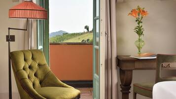 A room with a green armchair, a desk with a vase of flowers, and a view of a green landscape through a window.