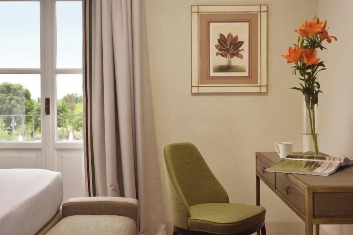 A hotel room with a bed, a chair, a desk with a vase of flowers, a framed picture on the wall, and a window with curtains.
