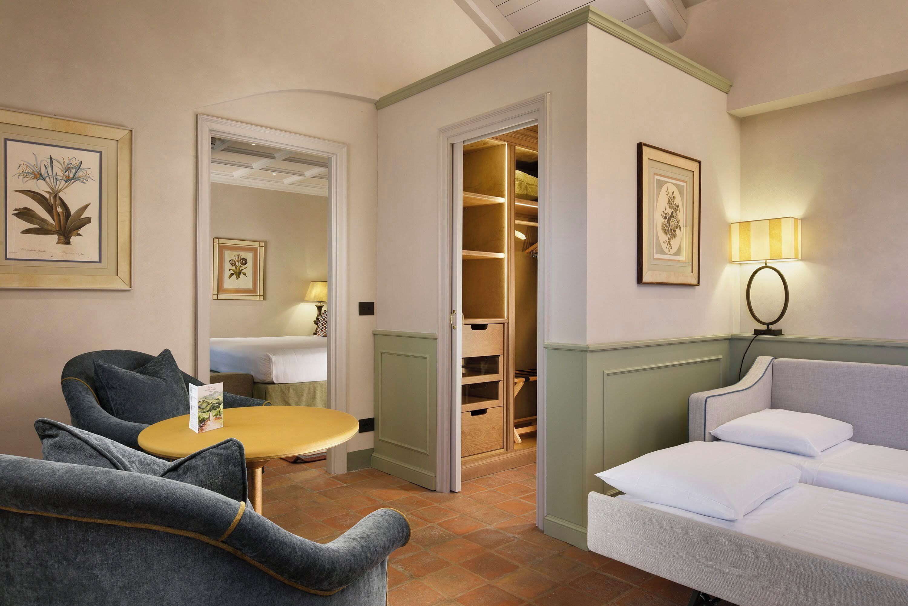 Quarto de hotel com sofá, mesa pequena, luminária, espelho e guarda-roupa.