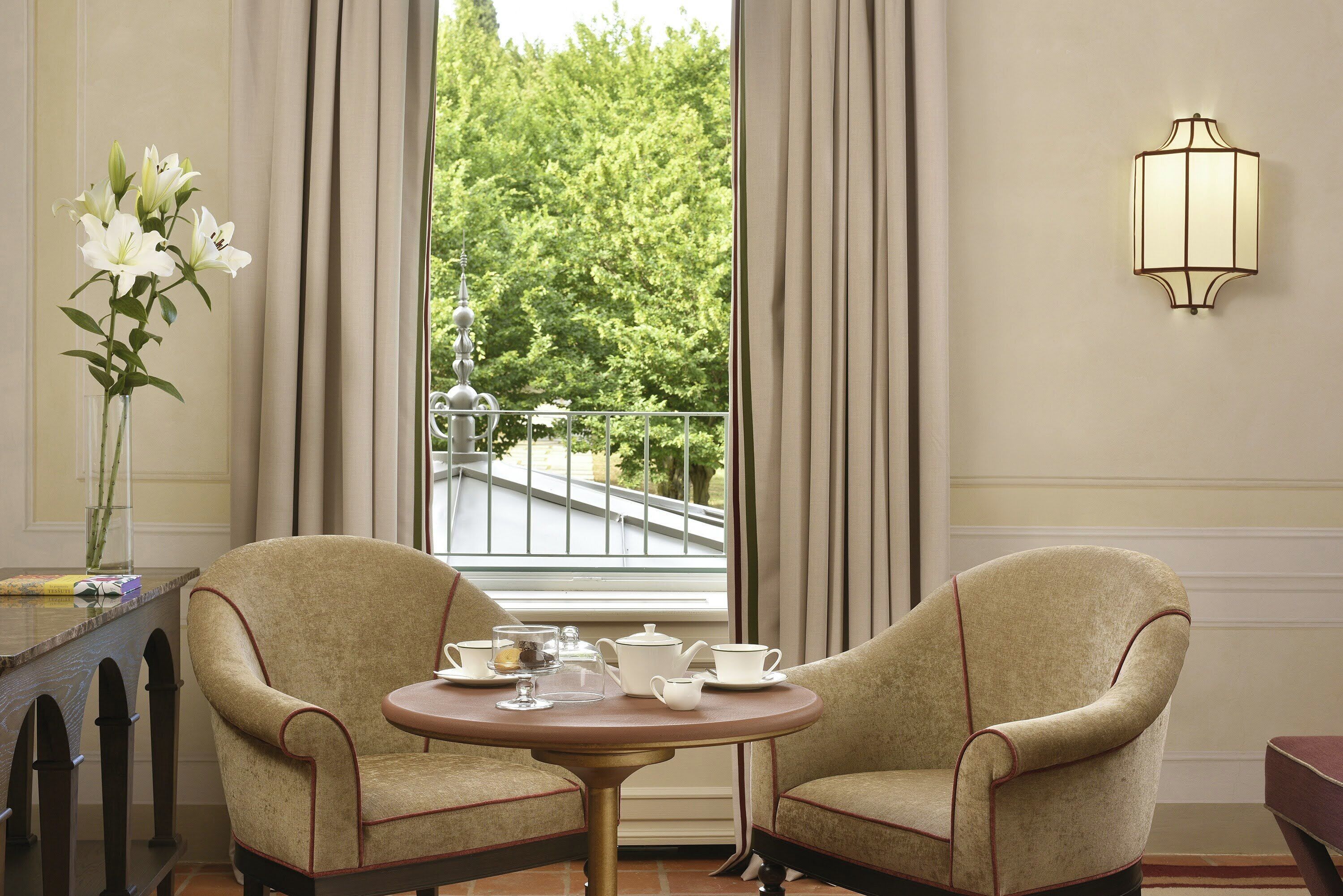 Une pièce comprenant deux fauteuils, une petite table ronde avec un service à thé, un vase de fleurs et une vue sur un balcon agrémenté de verdure.