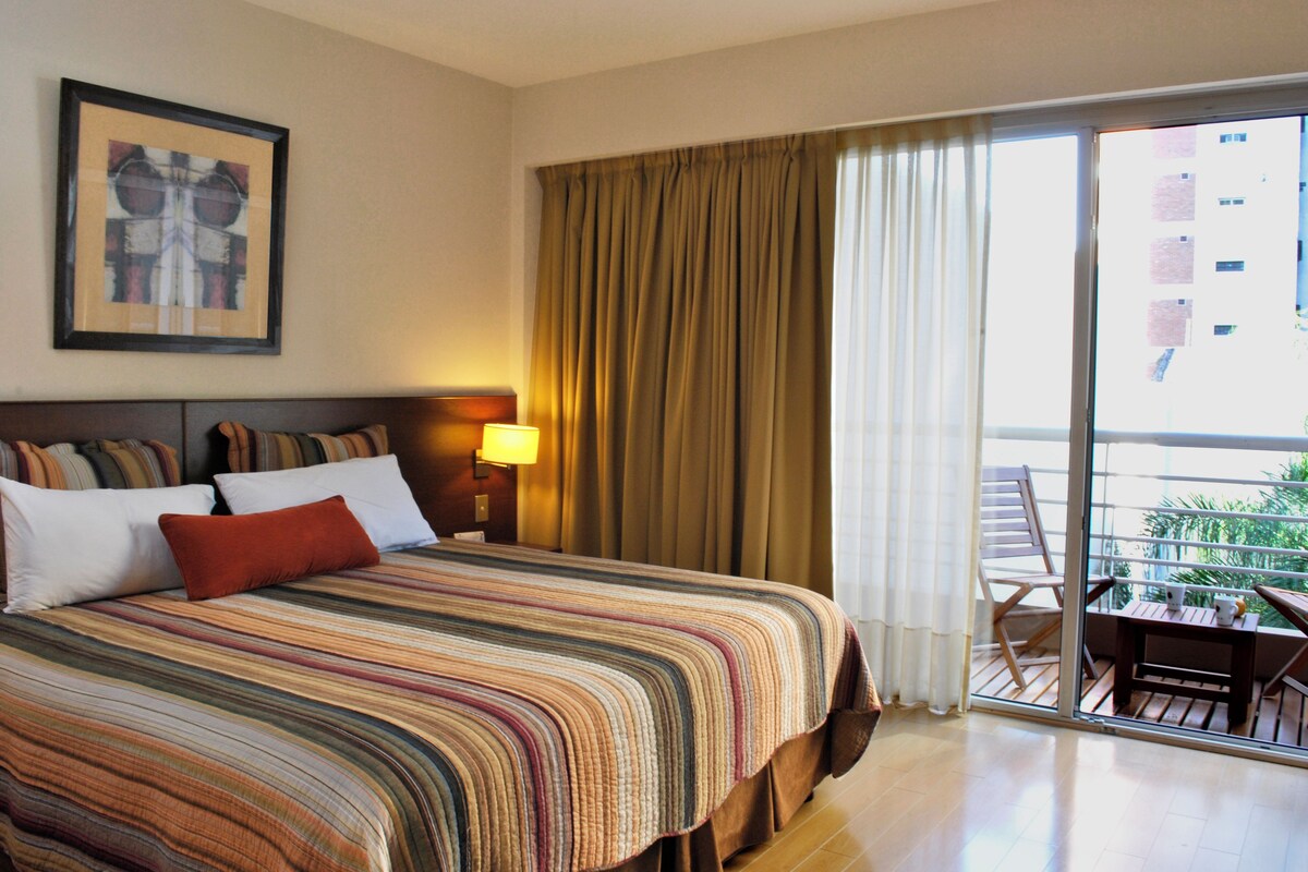 Habitación de hotel con cama, mesita de noche, lámpara, ventana con cortinas y balcón con silla y mesa.