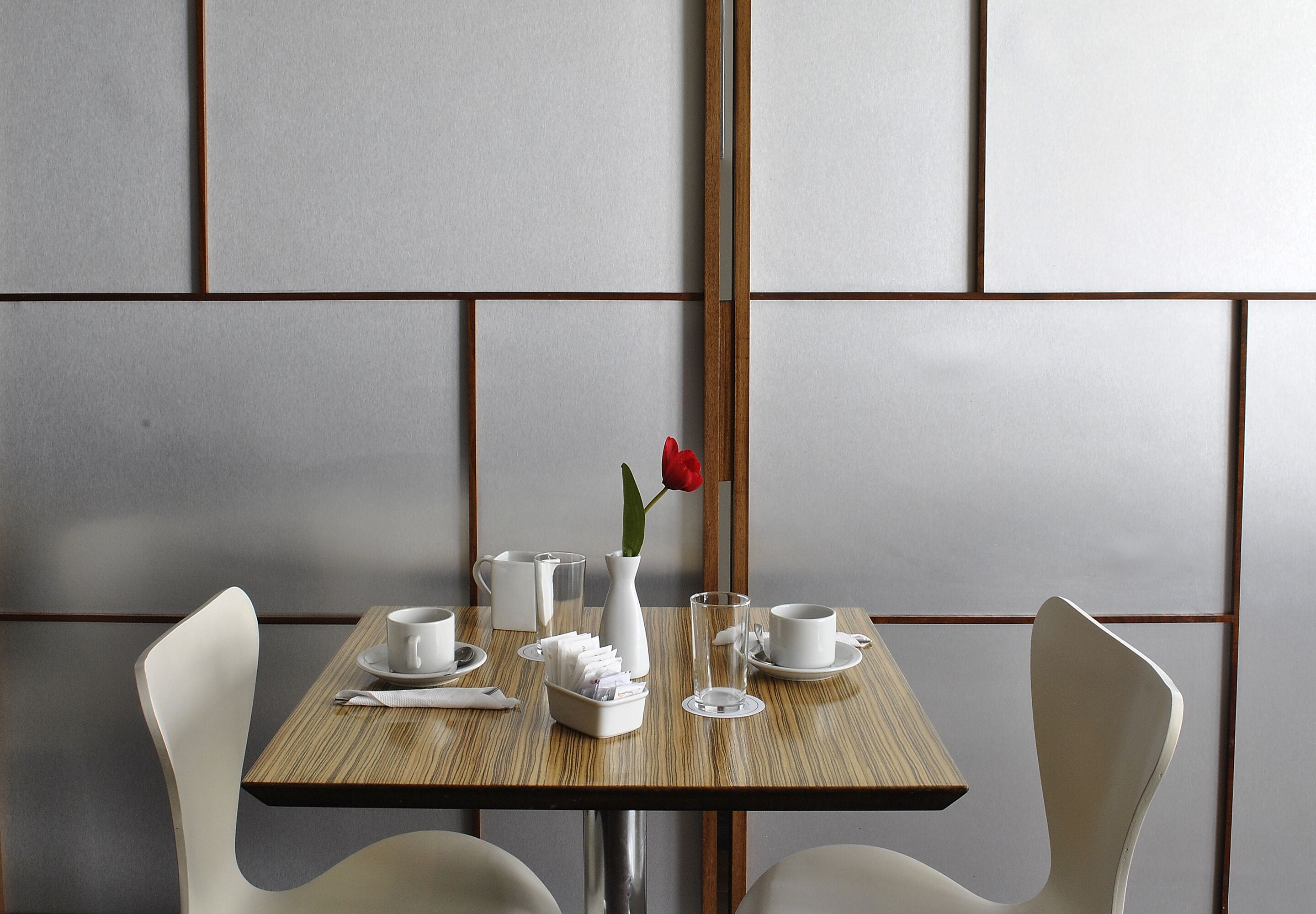 Una mesa de comedor con un florero de rosa roja, tazas blancas y una mesa de madera.