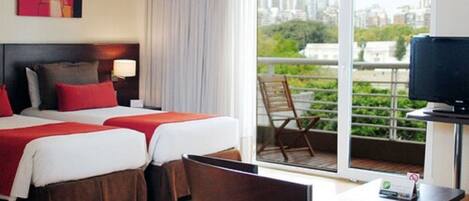 Habitación de hotel con dos camas, un escritorio, una silla, televisión y balcón con vista a la ciudad.