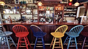 Una zona de bar con mostrador de madera, diferentes botellas, vasos y una variedad de sillas coloridas.