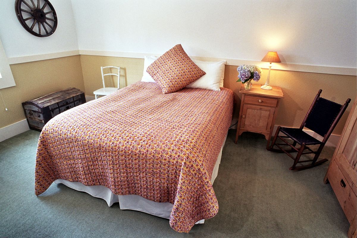 Una cama bien hecha con cubrecama y almohadas estampadas, una mesita de noche de madera con una lámpara y un florero, una mecedora y un pequeño cofre de madera.