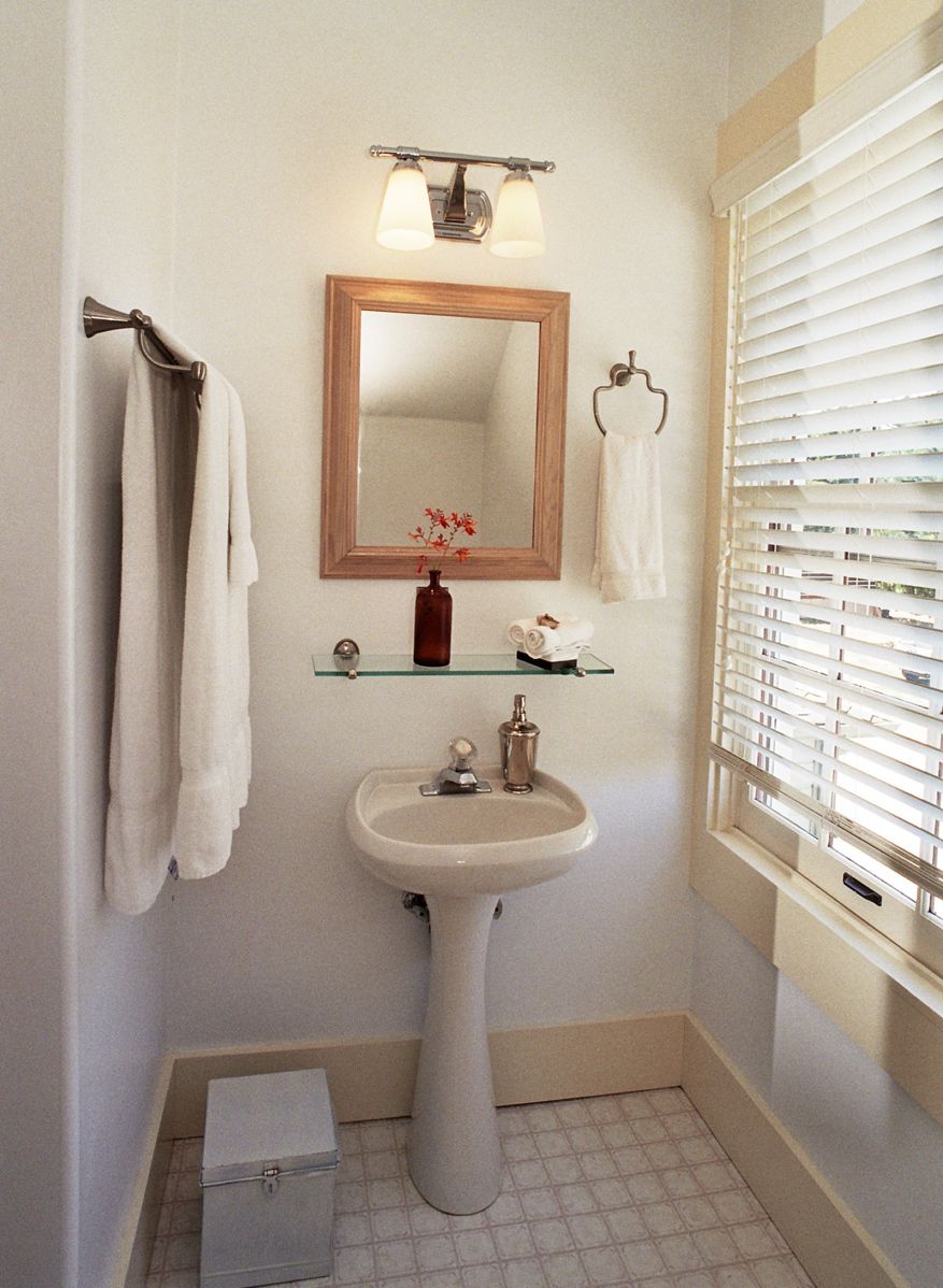 A bathroom with a pedestal sink, a mirror, a towel rack, a shelf with toiletries, and a window with blinds.