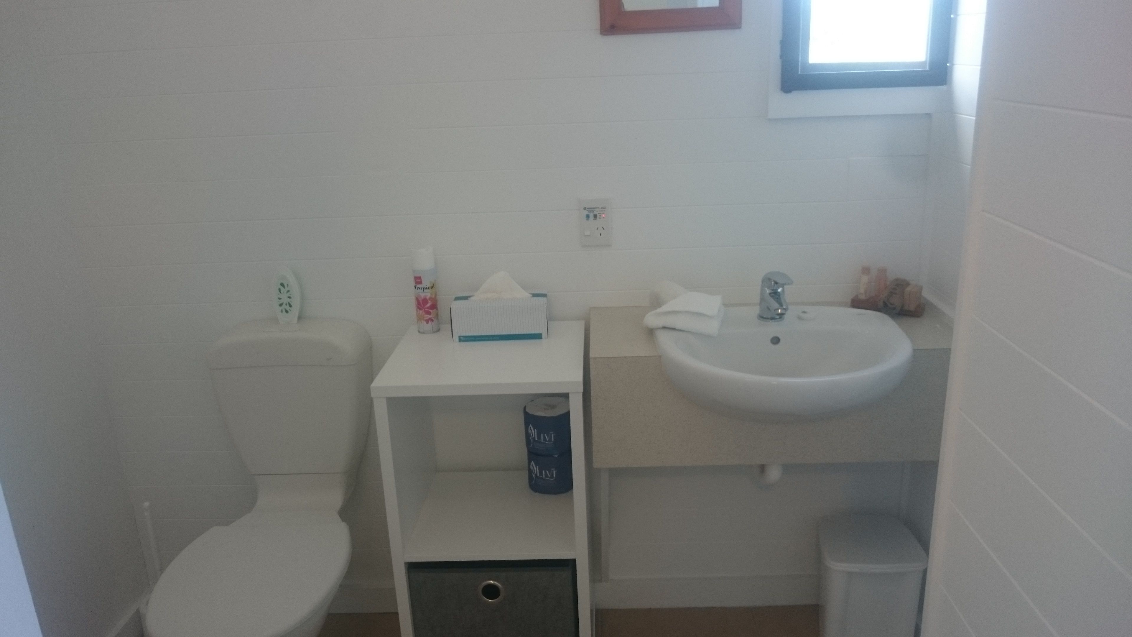 Un baño pequeño y limpio con inodoro, lavamanos y un estante con artículos de aseo.