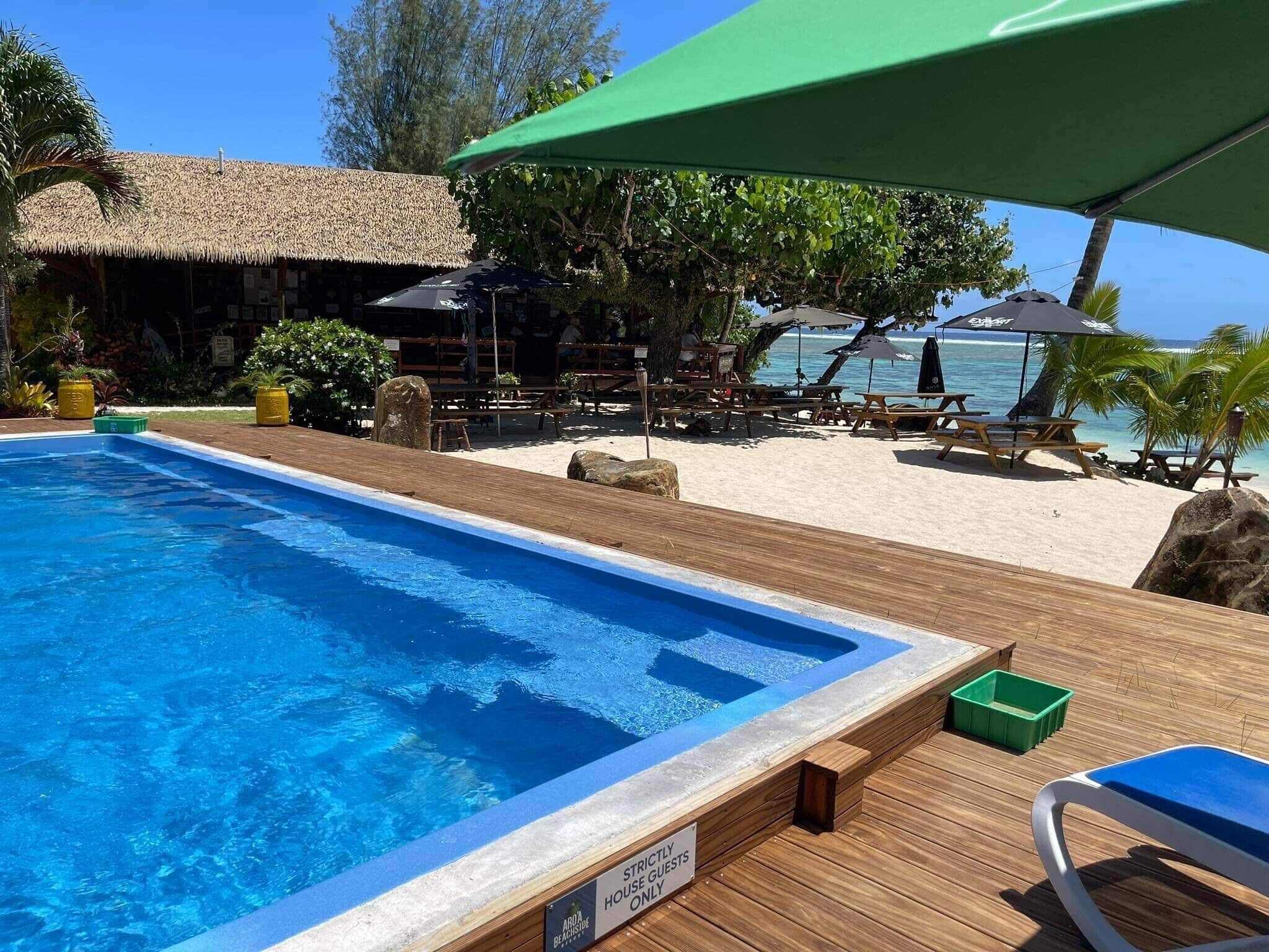 A pool area with a wooden deck, a sign indicating it is for strictly house guests only, and a beach with umbrellas and lounge chairs in the background.
