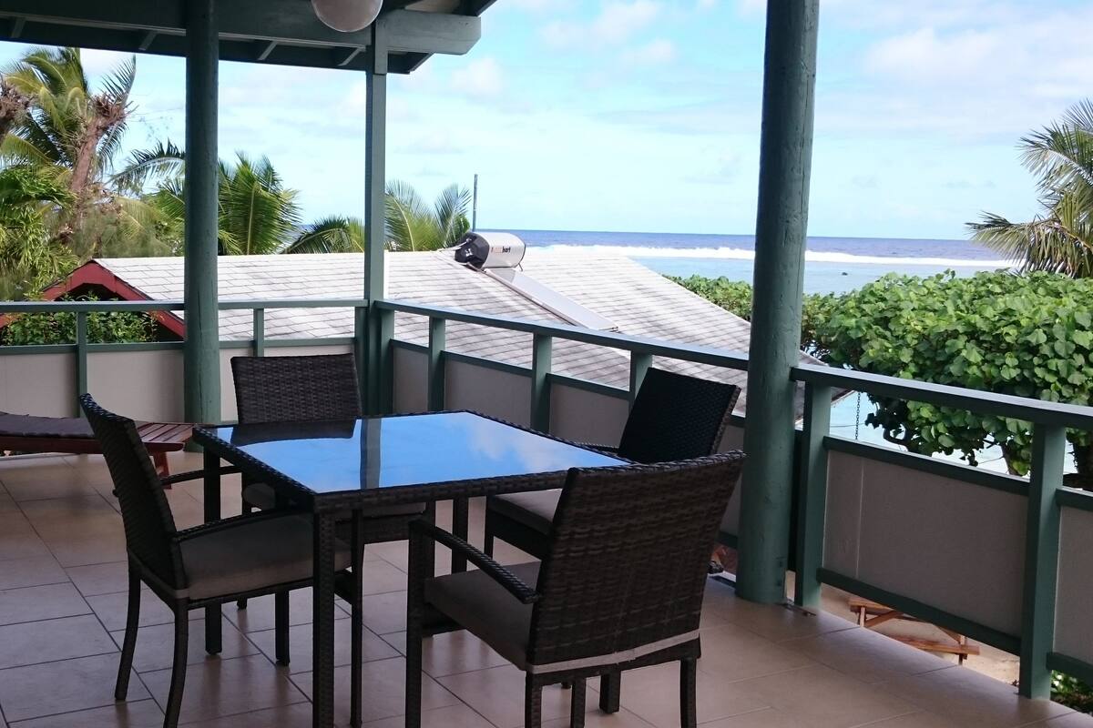 Un balcón con mesa y sillas, con vistas a una playa y palmeras.