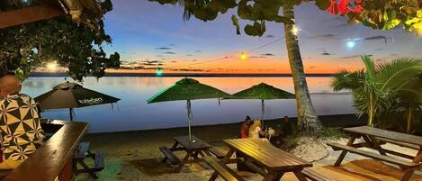 Un restaurant situé en bord de mer, avec une terrasse, une vue sur le coucher de soleil et un barbecue.
