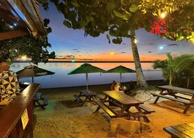 Un restaurante frente a la playa con mesas al aire libre, vista al atardecer sobre el agua y una parrilla.