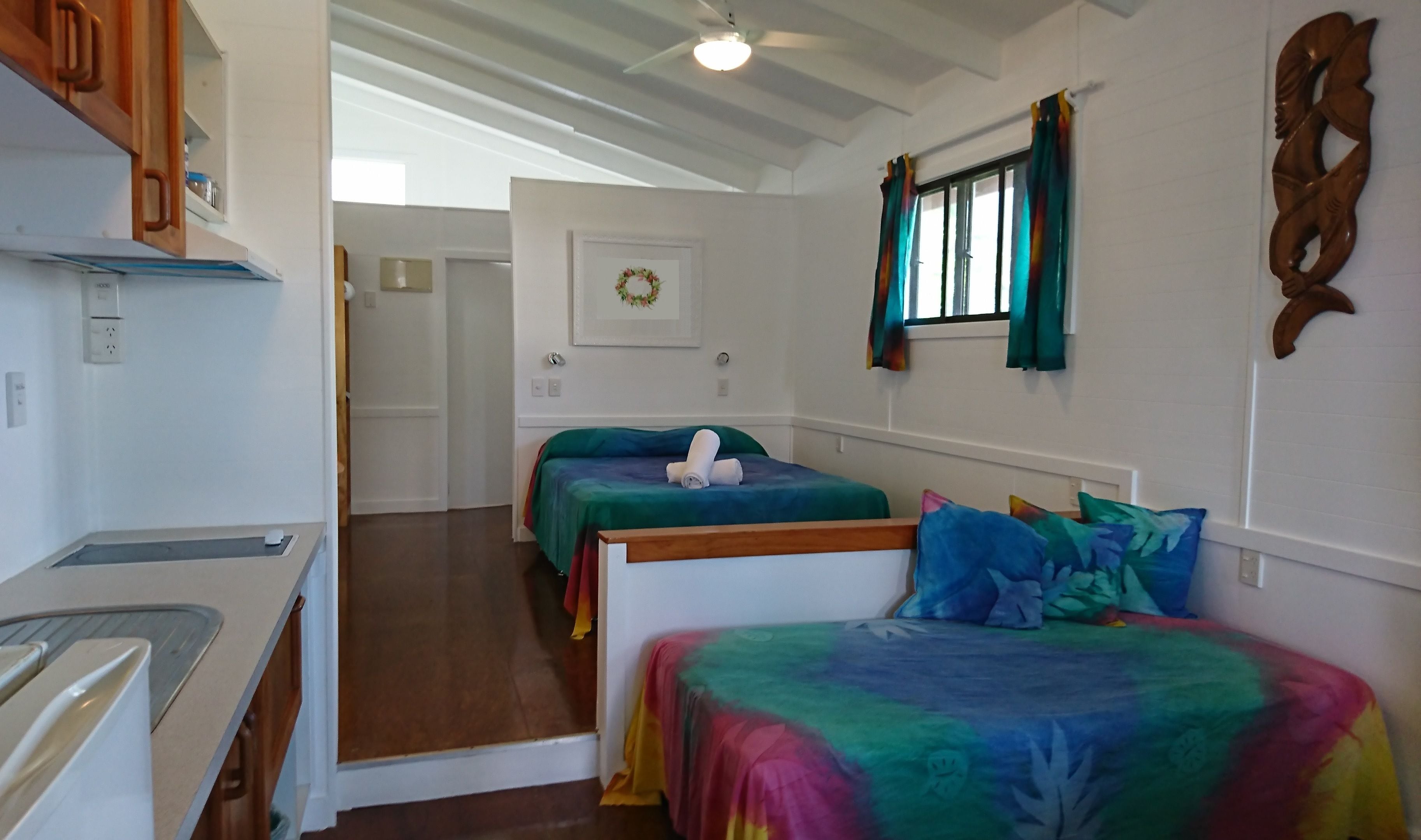 Ein Zimmer mit zwei Betten, einer Küchenzeile, einem Deckenventilator und einem Fenster mit Vorhängen.