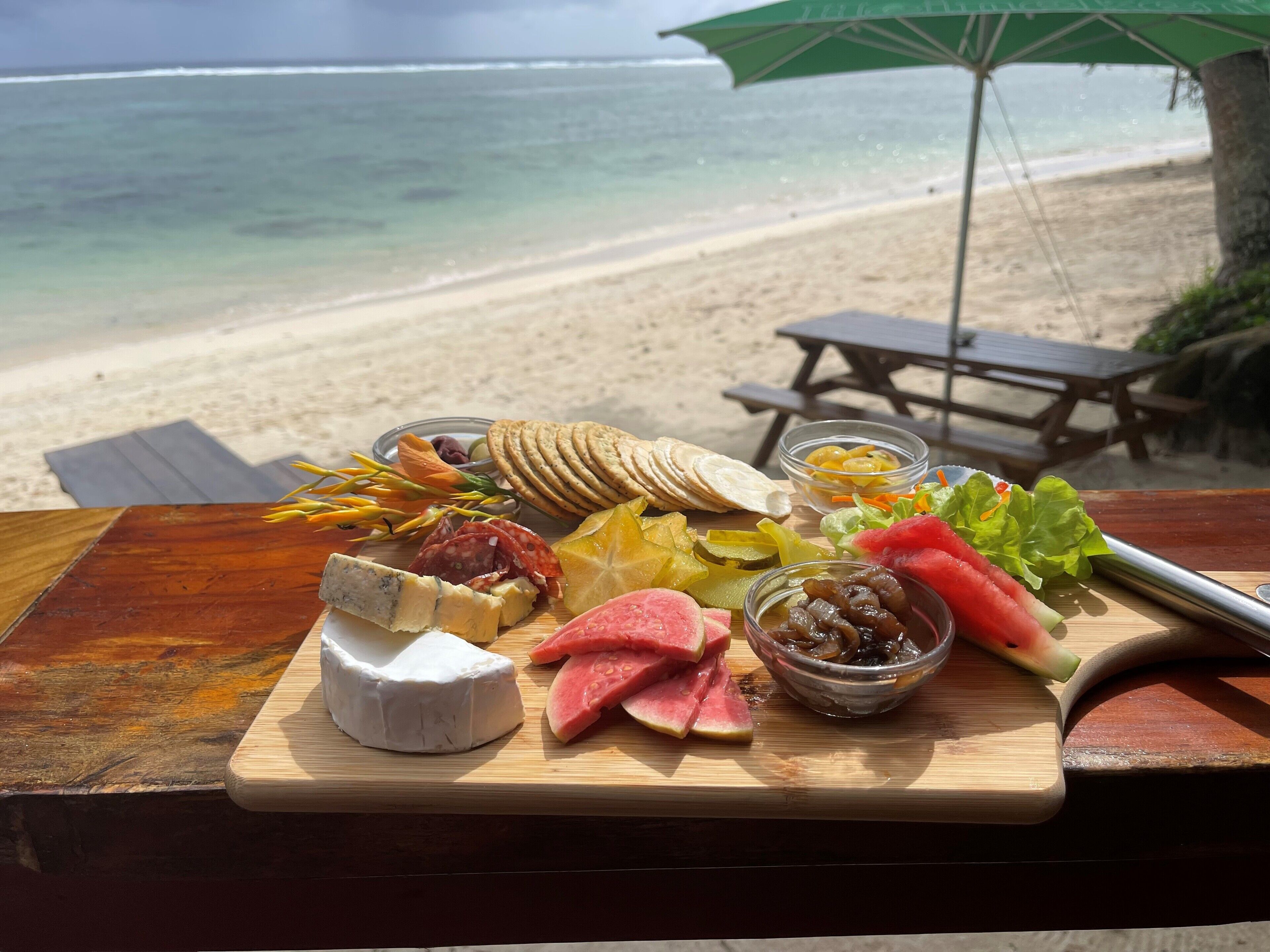 Una tabla de madera con diversos quesos, carnes y frutas en una playa con vista al océano.