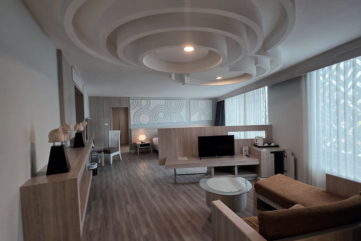 Una habitación de hotel moderna con piso de madera, televisor, sofá, una mesita y un ventanal grande con persianas.