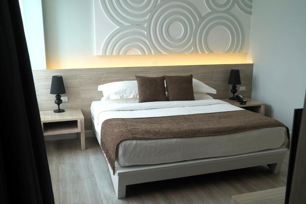 Quarto de hotel moderno com uma cama grande, duas mesas de cabeceira com abajures, um painel decorativo na parede e piso em tom claro.