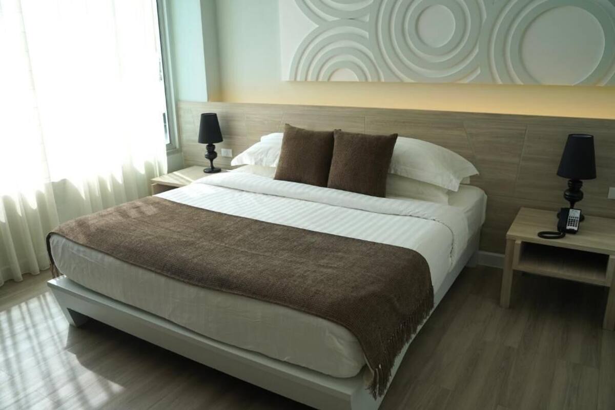 Una habitación de hotel moderna con una cama grande, dos mesitas de noche con lámparas, una pared con diseños circulares y una ventana con cortinas.