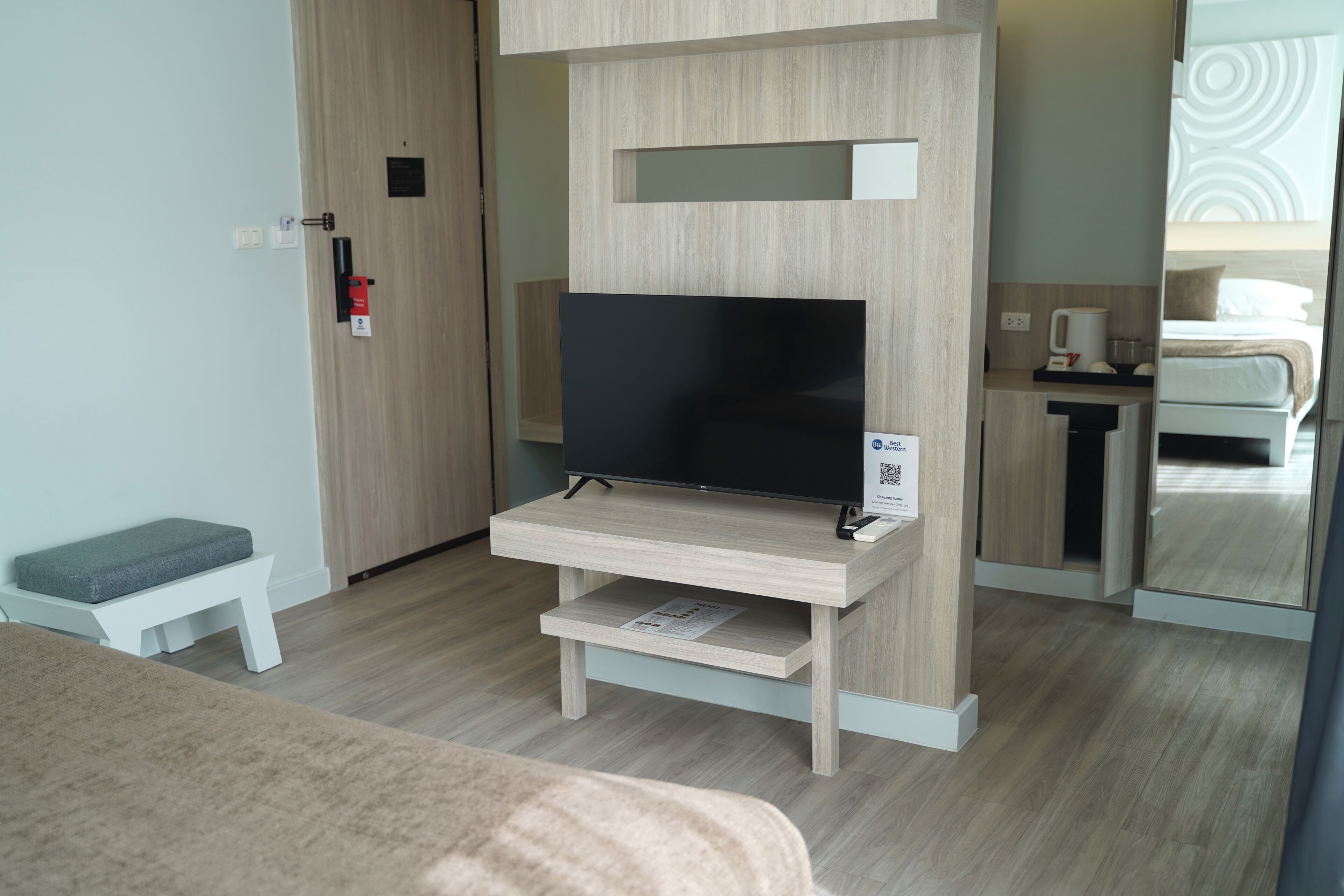 Una moderna camera d'hotel con una TV a schermo piatto su un supporto in legno, una piccola zona salotto e uno specchio che riflette l'ambiente.