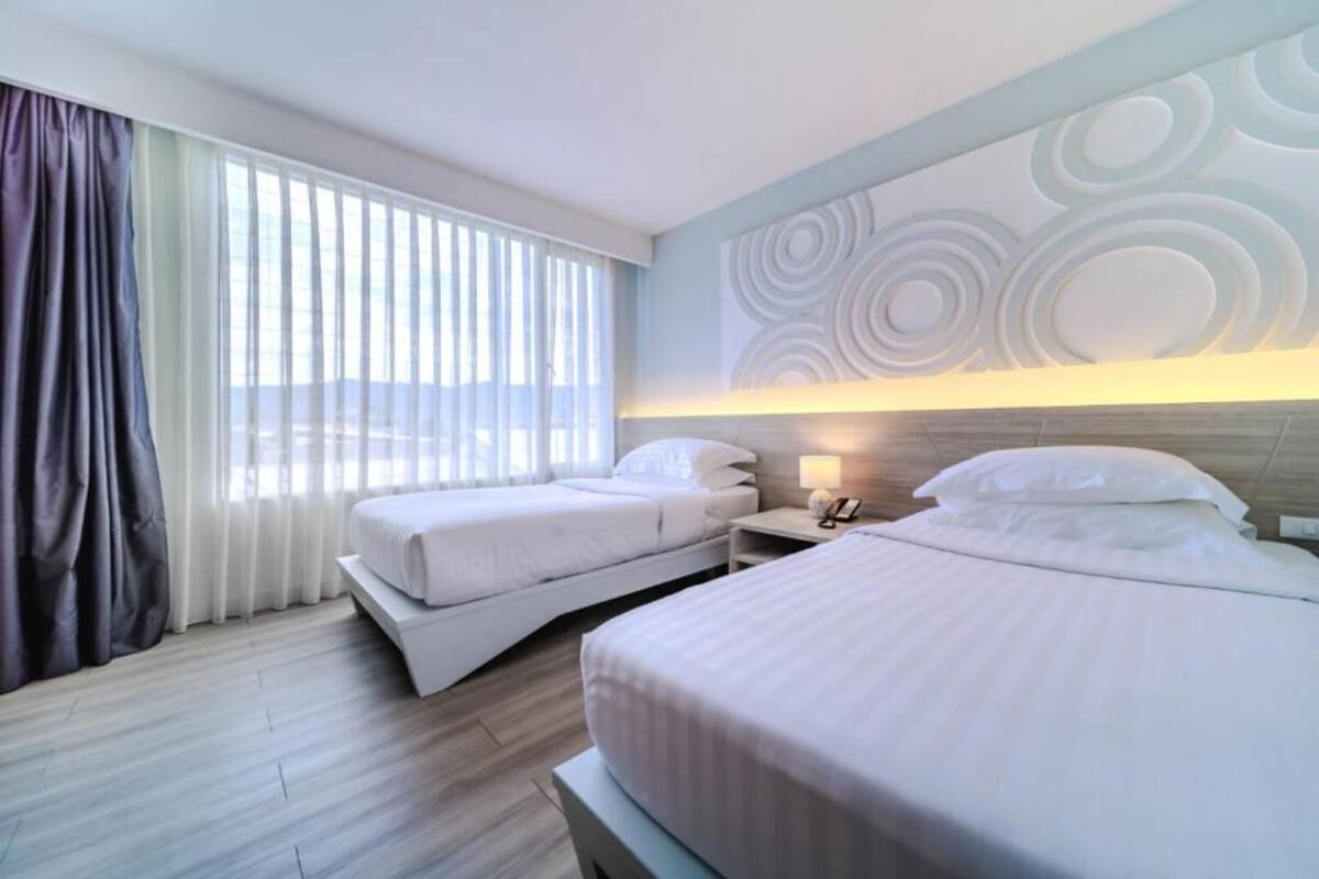 Habitación de hotel con dos camas, un ventanal con cortinas, una mesita de noche con lámpara y una pared con diseños circulares.