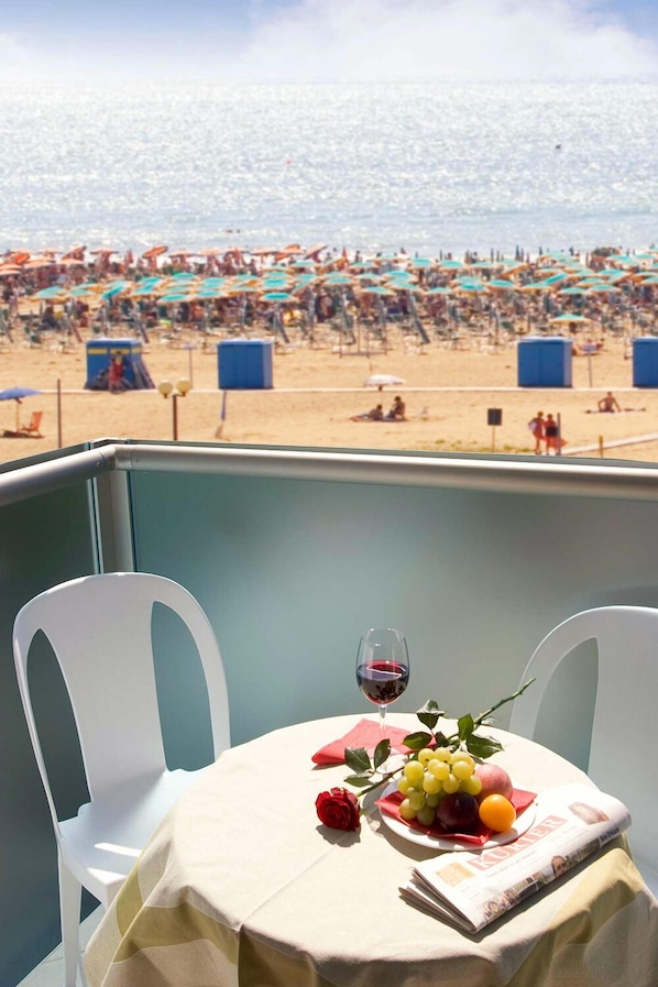 A table with a newspaper, a glass of red wine, and a fruit arrangement on a balcony overlooking a beach.