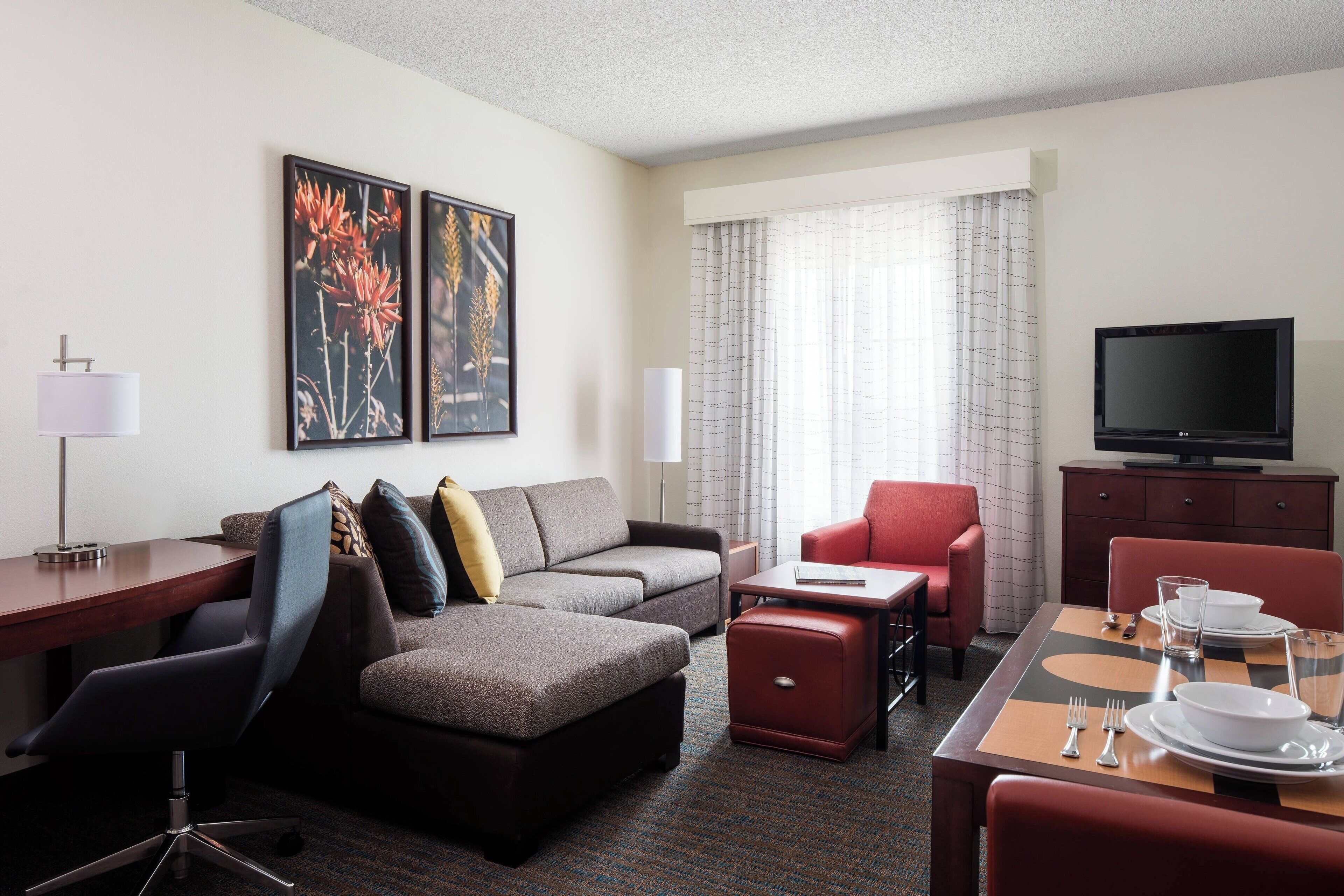 Ein Hotelzimmer mit Sofa, Sessel, Esstisch, Fernseher und Kunstwerk an der Wand.