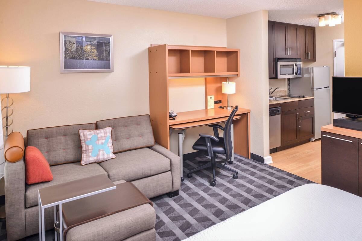 A hotel room with a sofa, a desk, a chair, a bed, a TV, a microwave, a refrigerator, and a small kitchen area.