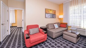 A hotel room with a red armchair, a grey sofa, a small table, and a lamp. There is a window with sheer curtains and a radiator.