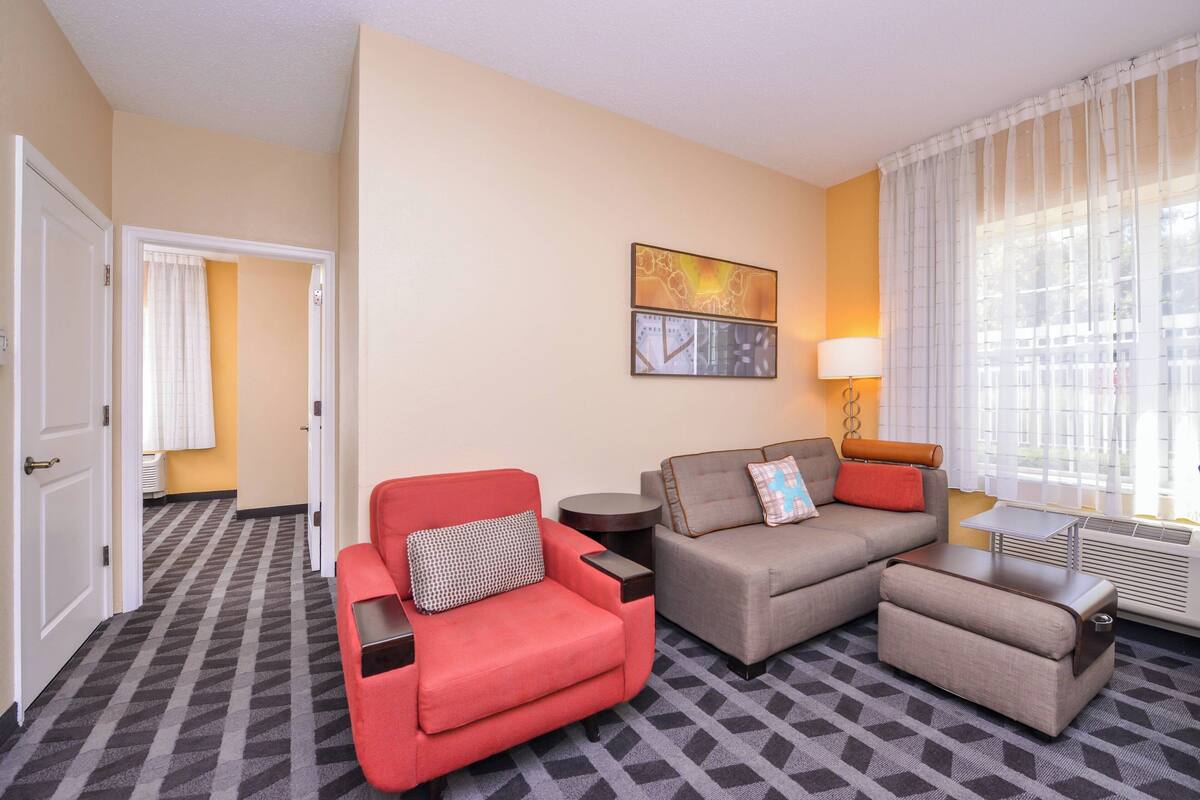 A hotel room with a red armchair, a grey sofa, a small table, and a lamp. There is a window with sheer curtains and a radiator.