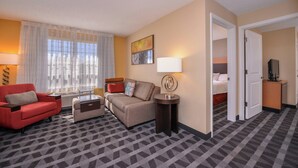 A hotel room with a sofa, armchair, and a small table. There is a bed in the adjacent room.