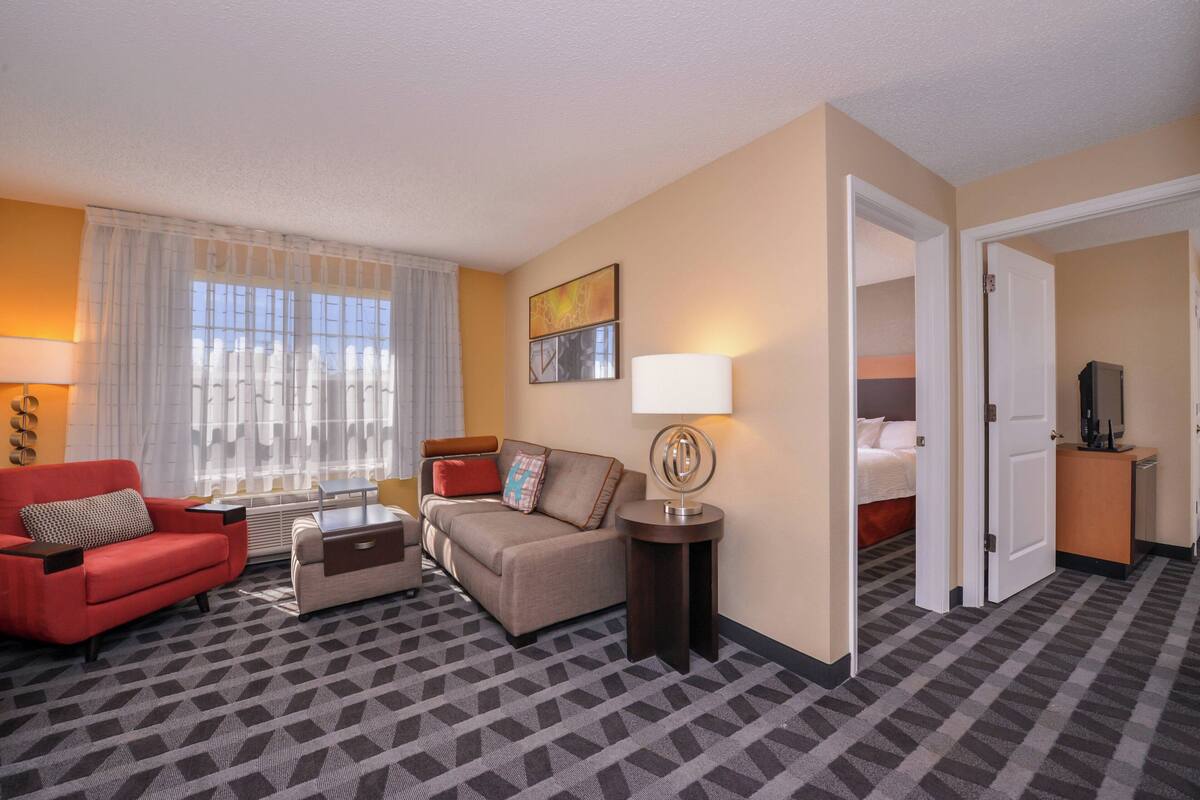A hotel room with a sofa, armchair, and a small table. There is a bed in the adjacent room.