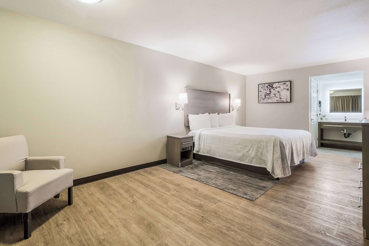 A hotel room with a bed, bedside table, chair, and a bathroom visible through an open door.