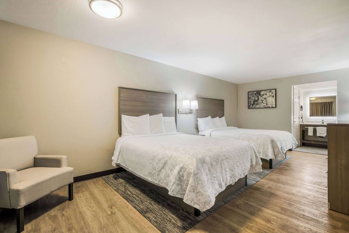 A hotel room with two beds, a sofa, a bathroom with a sink and mirror, and a wooden floor.