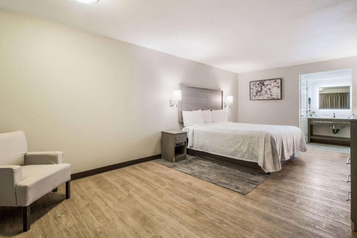 A hotel room with a bed, bedside table, chair, and a bathroom with a mirror and sink.